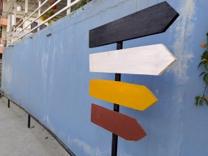 A wooden signpost with four colored arrows—black at the top, followed by white, yellow, and red—mounted on a black stand. 