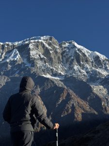 A person wearing a dark jacket and hood stands on a mountainous landscape, gazing at snow-capped peaks under a clear blue sky. 