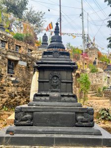 A black, ornate stone stupa with intricate carvings stands in front of a stone wall lined with Buddha statues and colorful prayer flags.