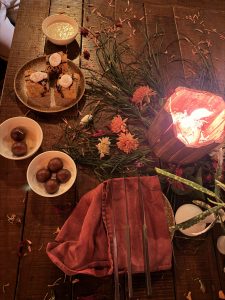 A rustic wooden table with sweets, a creamy dish, cake, skewers on a red cloth, a glowing lantern, and scattered flower petals.