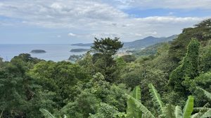 An aerial view of a tropical coastline with green forests, distant islands, and a blue sea under cloudy skies.