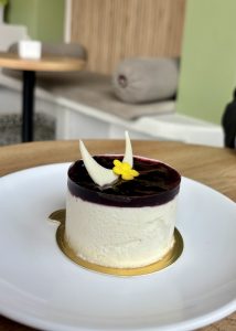 
A close-up view of a small, round cheesecake topped with a layer of dark berry glaze. It features two delicate white chocolate decorations resembling horns, and a tiny yellow edible flower is placed on top.