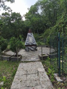 A stone monument with a red brick top is situated in a small, fenced area surrounded by greenery. The ground is paved with stone slabs, and a walking path leads towards the monument.