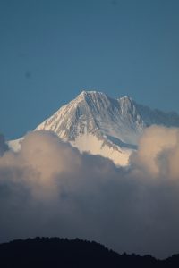 A tall, snow-capped mountain peak rises majestically against a blue sky, partially obscured by fluffy white clouds at its base. 