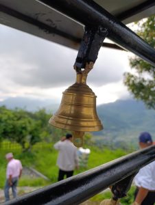 A close-up view of a brass bell hanging from a black metal structure, with a blurred background depicting greenery and a cloudy sky. 