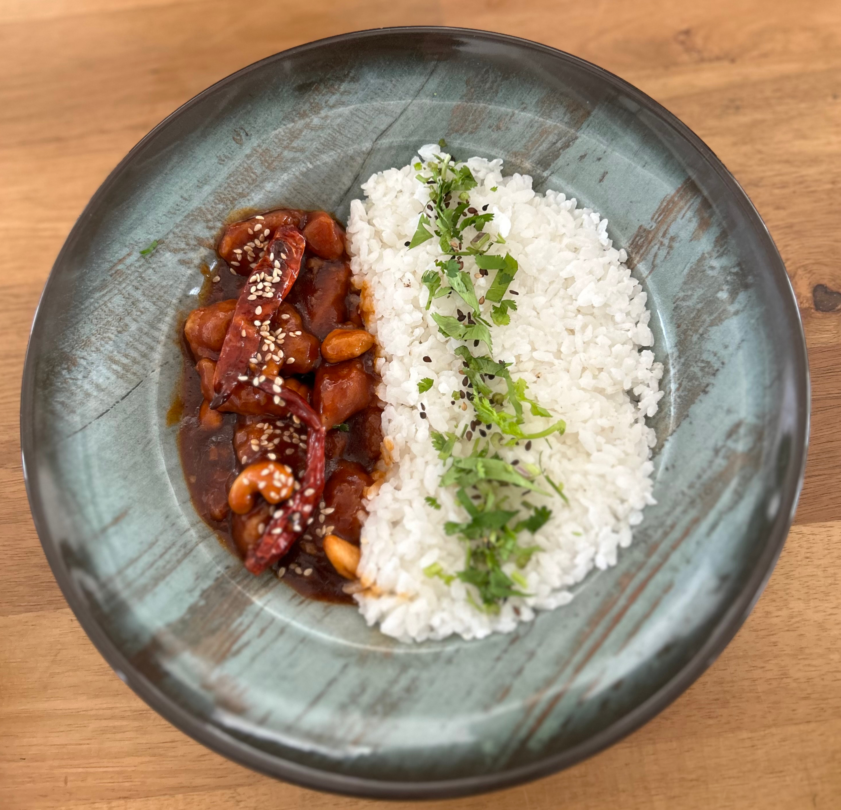 A round teal plate shows white rice with green herbs on one side and a saucy chicken dish with peanuts, dried red chilies, and sesame seeds on the other, set on a wooden table
