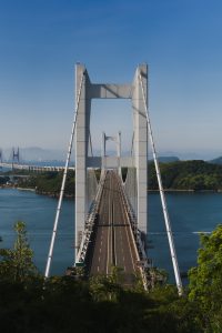 A frontal view of the Seto Ohashi Bridge spanning the Seto Inland Sea. The bridge’s piers and suspension structure repeat into the distance, with the roadway stretching forward, framed by blue water, green islands, and a clear sky.
