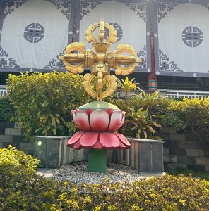 A large decorative structure stands in a garden, featuring a vibrant red and pink lotus flower base with broad petals.
