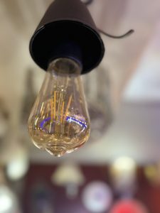 A close-up view of a vintage-style light bulb hanging from a black fixture. The bulb is transparent with a warm golden glow, showcasing intricate filament designs inside
