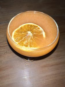 An orange-colored cocktail in a coupe glass is garnished with a dried orange slice and sits on a wooden surface.