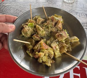A hand is holding a metallic plate filled with skewered pieces of grilled meat and vegetables, garnished with green herbs.