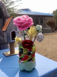 
A colorful arrangement of artificial flowers, including a large pink crocheted rose, a red crocheted rose, and yellow paper flowers, is displayed in a decorated plastic bottle wrapped in colorful tape