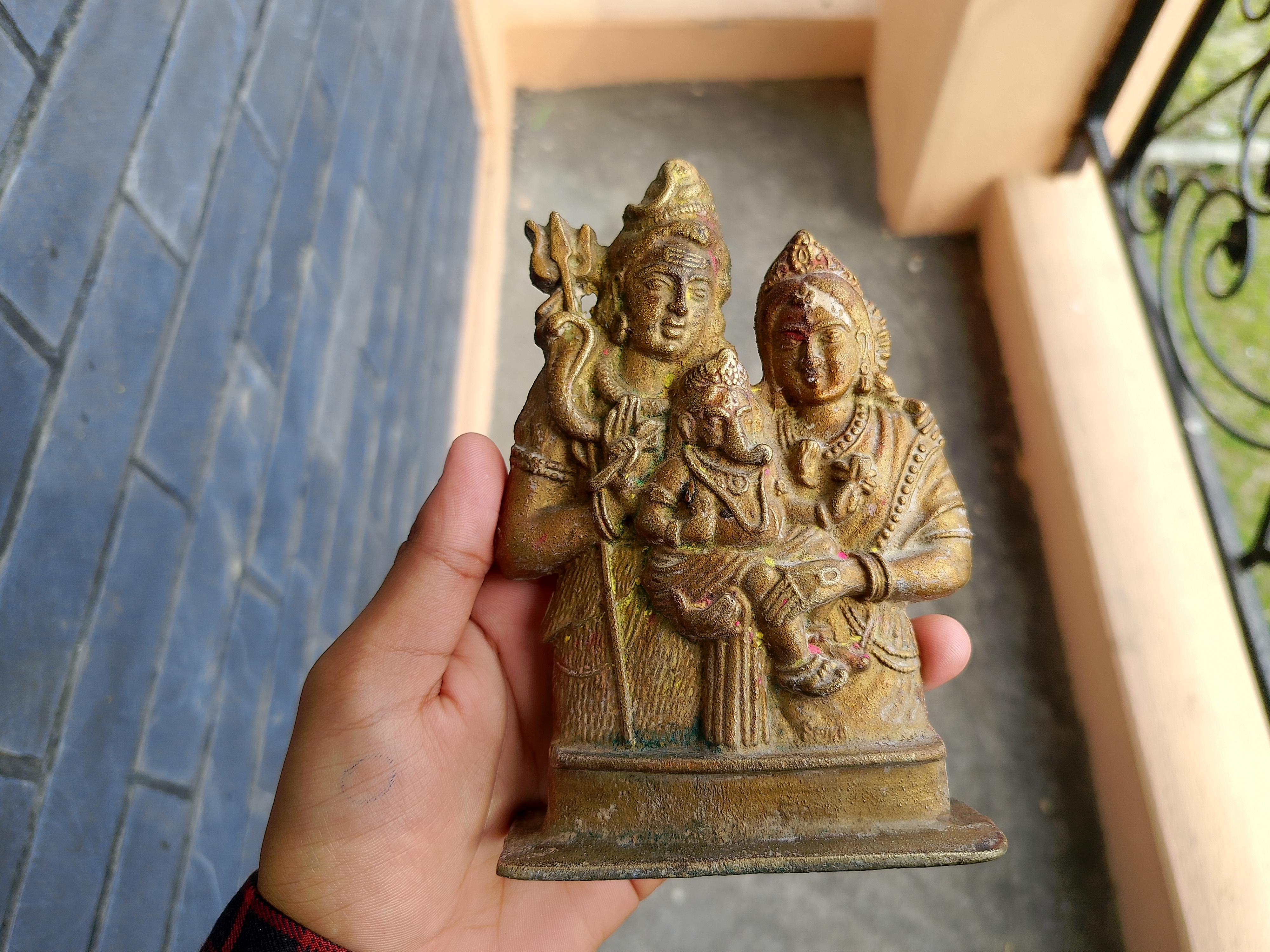 The image shows a small decorative statue being held in a person's hand. The statue features two figures, which may represent deities. One figure on the left has multiple arms, holding various objects, while the other figure on the right is adorned with intricate attire and jewelry. Both figures have expressive faces and a sense of serenity. The st
