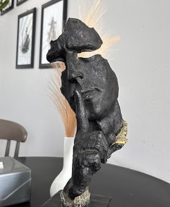 A black textured sculpture of a face with a finger held to the lips, symbolizing silence.