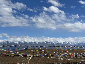 A panoramic view showcasing a vast landscape with colorful prayer flags fluttering in the foreground. In the background, majestic snow-capped mountains rise under a bright blue sky adorned with fluffy white clouds.