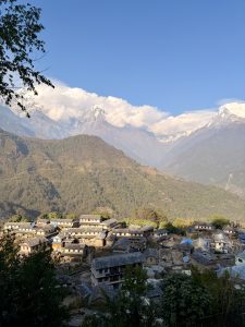 A panoramic view of snow-capped mountains under a clear blue sky, with Ghandruk village nestled among green terraced slopes in the foreground.