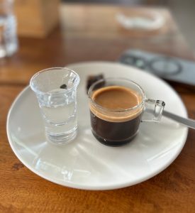 
A clear glass of water and a small glass cup of espresso sit on a white plate, accompanied by a small piece of dark chocolate, all placed on a wooden table