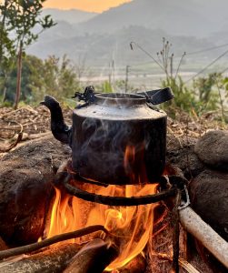 
A black kettle is perched over a vibrant fire made of logs and sticks, with smoke rising from its spout.