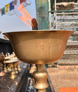 A close-up view of a brass offering bowl with a flame rising from its center, creating wisps of smoke. 