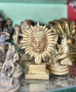A collection of ornate brass figurines is displayed, with a prominent golden sun-faced deity sculpture in the foreground. 