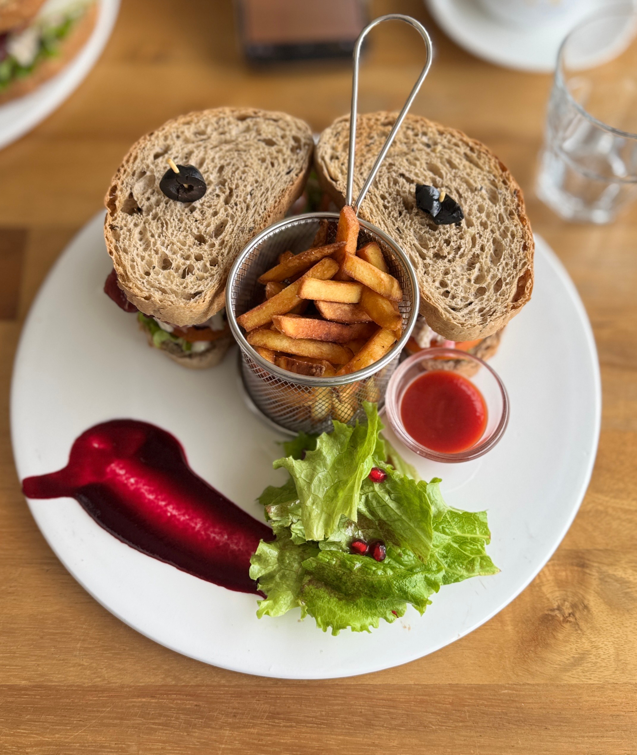 A white plate features a large sandwich made with two slices of brown bread, stuffed with various fillings and topped with a black olive. Nestled between the slices is a small metal basket filled with golden, crispy French fries.