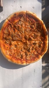 A freshly baked pizza sits on a white table. The pizza has a golden crust and is topped with a rich red tomato sauce, melted cheese, and slices of various meats, including sausage and pepperoni. Sunlight highlights the toppings, giving the pizza a glossy appearance.