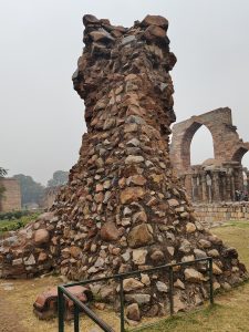 A weathered stone structure, featuring a mix of large and small sandstone pieces, stands prominently against a gray, overcast sky. In the background, remnants of arches and pillars can be seen, part of an ancient architectural site.