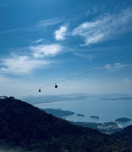 A scenic view from a mountain overlooking a body of water, with several small islands visible in the distance. Two gondolas are suspended on a cable, traversing the sky. Langkawi SkyCab, in Malaysia.
