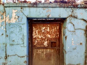 A weathered blue wall with peeling paint surrounds a rusty metal door. 