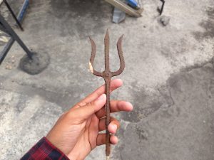 A hand is holding a small, rusty garden tool resembling a pitchfork with three pointed tines. The tool shows signs of age and wear, with a textured, uneven surface.