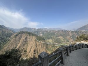 A wide view of rolling mountains under a clear blue sky, with green slopes, terraced fields, a winding road, and a distant suspension bridge.
