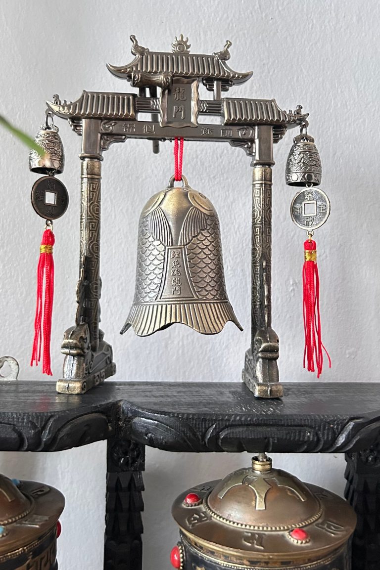 An ornate metal archway with a fish-shaped bell at the center, and smaller bells and coins hanging on red cords.