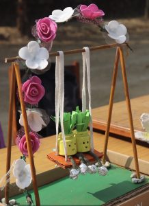 A colorful craft display featuring a small swing made from bamboo sticks, decorated with pink and white artificial flowers at the top. 