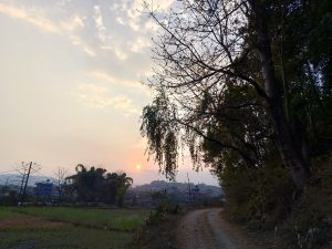 A serene sunset landscape with a winding dirt path, sparse trees, distant hills, and silhouetted buildings.