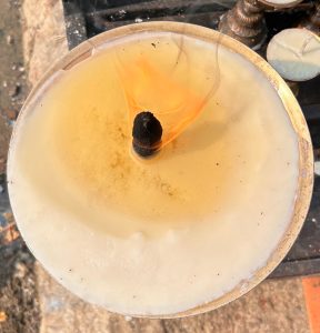 A close-up view of a candle with a burning wick, which is surrounded by a pool of melted wax. 