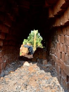 
A view through a brick archway, leading out to a garden with banana plants visible in the background. The arch is made of red bricks, with some bricks appearing cracked or weathered