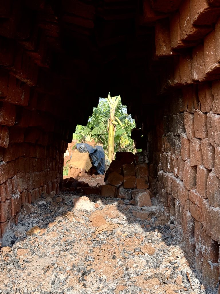 A view through a brick archway, leading out to a garden with banana plants visible in the background. The arch is made of red bricks, with some bricks appearing cracked or weathered