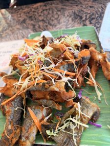 A plate of crispy fried fish garnished with shredded cabbage and carrot, served on a banana leaf, set on a patterned table surface.