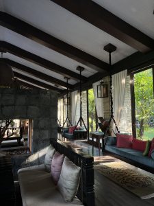 A cozy indoor lounge area featuring high ceilings with dark wooden beams. The space is illuminated by natural light streaming through large windows adorned with sheer white curtains. Several seating arrangements include long, cushioned benches with pink and green pillows and a dark wooden railing. 