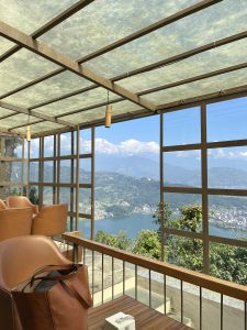 A cozy seating area with brown chairs and a wooden table holding a stylish bag, overlooking a serene lake and mountains.