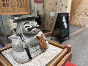 Detail of a polished stone Ebisu statue with a carved smile and sea bream, set within a Japanese covered shopping arcade.
