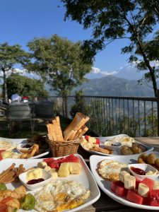 A picturesque outdoor breakfast setting featuring several plates of food. The foreground showcases two plates containing sunny-side-up eggs, fresh fruits like watermelon and banana, and a small dish with ketchup and butter. A basket of breadsticks is also visible. 