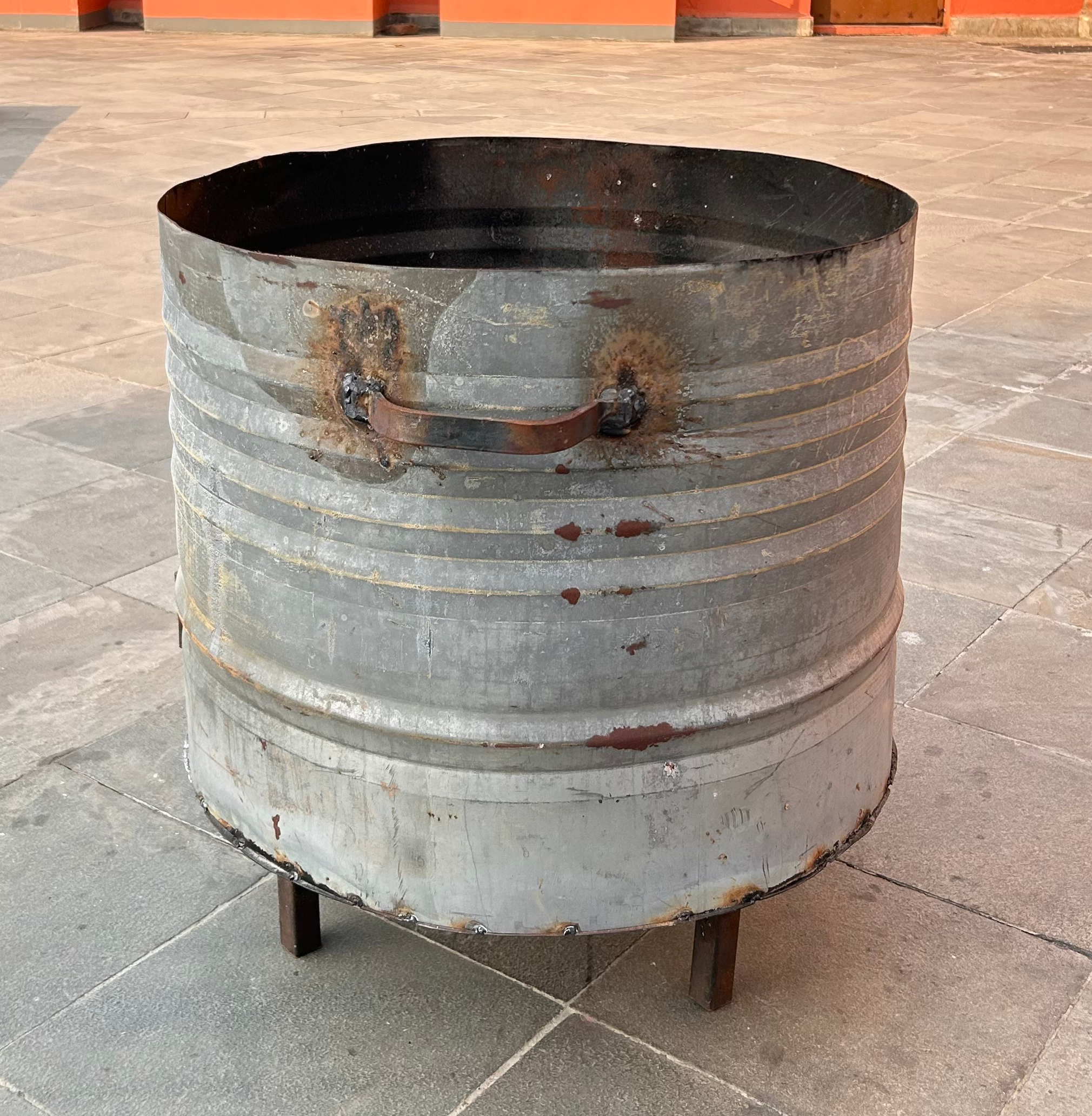 A large, round metal barrel with a slightly rusted surface, open rim, side handle, and four small legs, set on a stone floor.