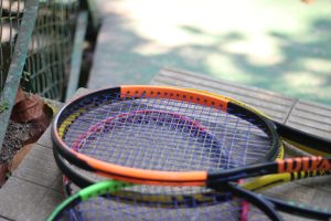A close-up of several tennis rackets stacked on a textured surface, showing colors like orange, red, green, and black.
