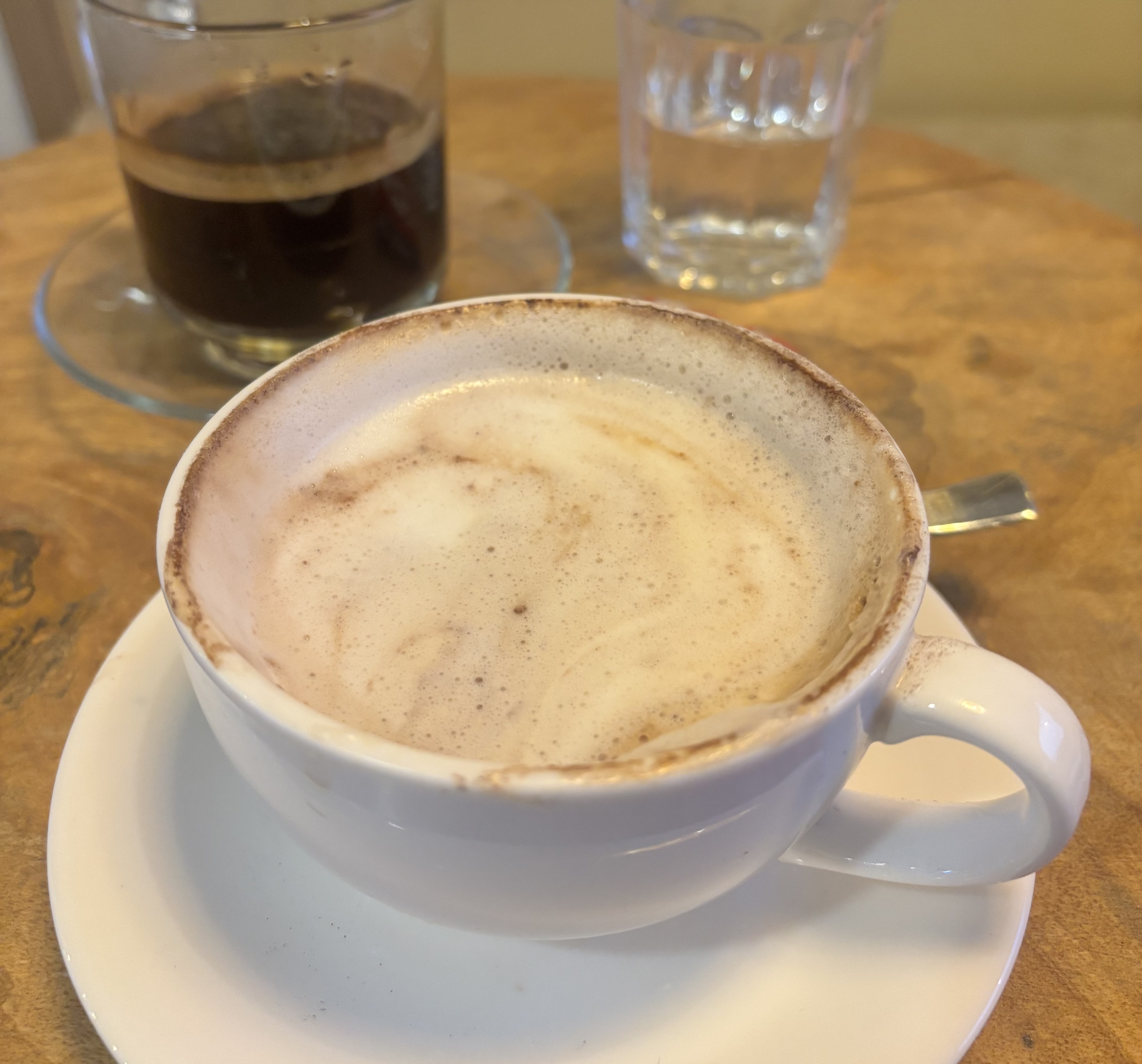 A close-up view of a white coffee cup filled with frothy coffee, resting on a small white saucer.