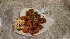 A white, heart-shaped plate holding a serving of crispy, fried chicken pieces coated in a dark, flavorful sauce, garnished with fresh green herbs.