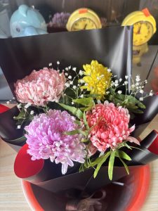 A bouquet of vibrant flowers arranged in a black paper wrap, featuring pink, purple, coral, and yellow blooms, accented with delicate white baby's breath.