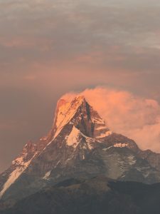 A majestic Fishtail (Machapuchare) mountain peak, partially covered in snow, is illuminated by a warm orange glow from the setting sun. 