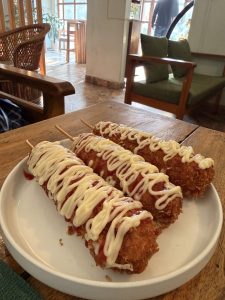 A white plate holds three deep-fried corn dogs skewered on wooden sticks, each drizzled with mayonnaise and ketchup in a decorative pattern.