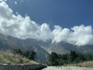
A scenic view of a mountainous landscape under a partly cloudy sky. The foreground features a narrow road bordered by a stone wall, with lush greenery and trees on the left side